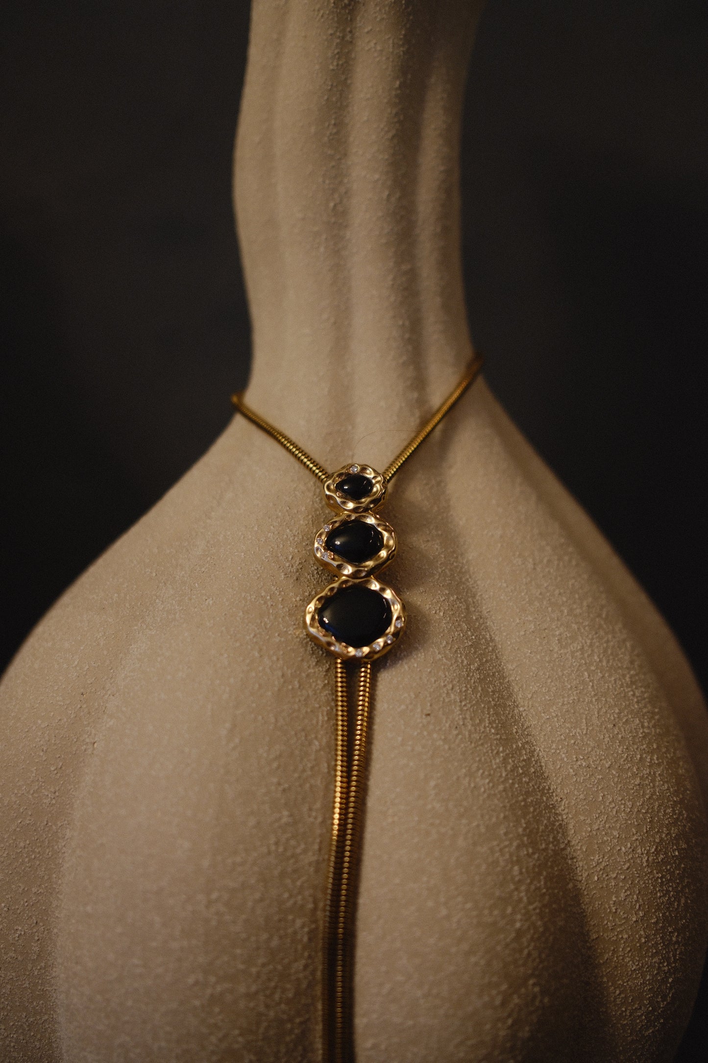 BOLOLA The Merriam necklace in onyx gold plated with lab diamonds adorning the stone. The necklace is draped over a curved beige vase. 